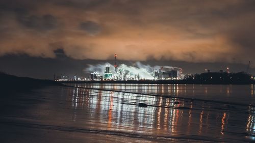 Illuminated factory by river against cloudy sky at night