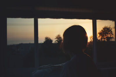 Silhouette woman looking through window at sunset