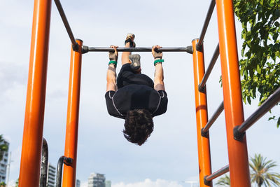 From below anonymous male athlete hanging upside down on bar against cloudy sky during fitness workout on sports ground in city