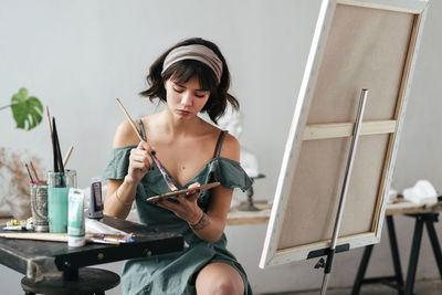 Young woman painting on canvas while sitting on table