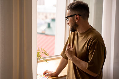 Side view of young artist man standing by window at home