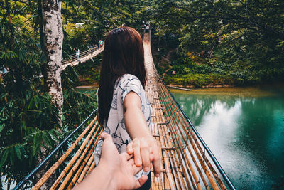 Cropped hand of man holding woman on bamboo footbridge over river in forest