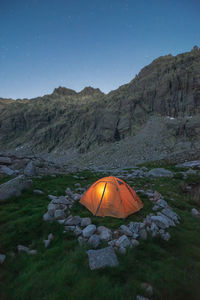 Picturesque view of tent on moss with stones against rugged mount under blue sky in twilight