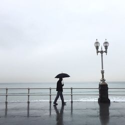 Side view of man walking under umbrella during rainfall by sea against sky