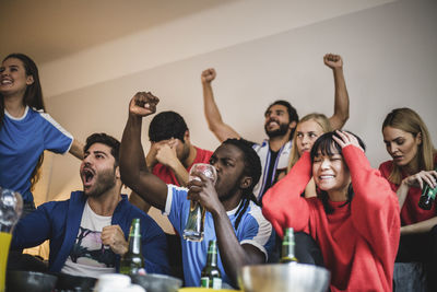 Multi-ethnic friends watching soccer match while sitting against wall at home