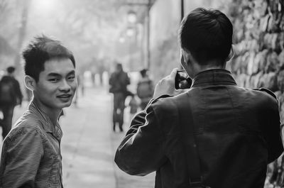 Portrait of man standing with friend photographing through mobile phone by wall