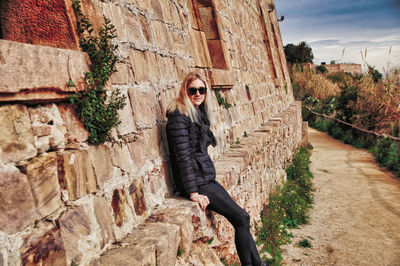 Portrait of woman wearing sunglasses while sitting on stone wall