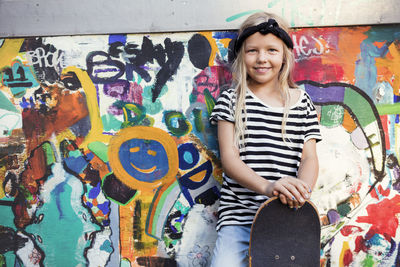 Portrait of smiling girl holding skateboard while standing against graffiti wall