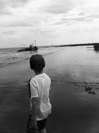 Rear view of boy walking on shore against cloudy sky