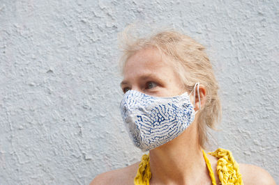 Calm middle aged female wearing protective mask standing on street near concrete building and looking away during coronavirus epidemic