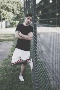 Portrait of man with arms crossed while leaning on chainlink fence at field