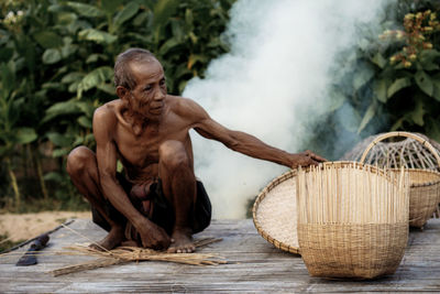 Full length of shirtless man making wicker basket 