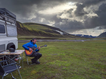 Man playing guitar while relaxing by off-road vehicle against cloudy sky during sunset