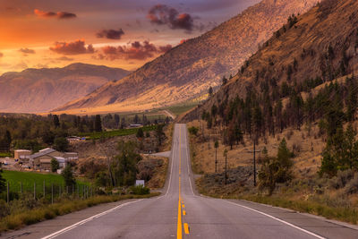 Country road by mountains against sky during sunset