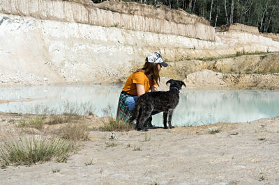 Young woman hiker in cap sitting on sandy shore of blue lake with fluffy dog, travel with pets