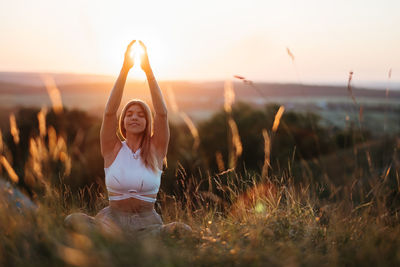 Young woman sitting in meditation yoga pose and catching sun by hands at sunset	
