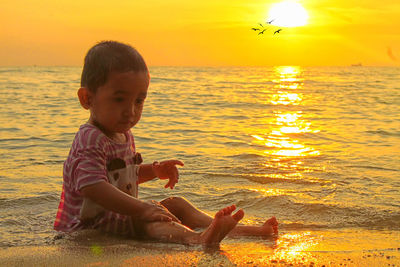 Cute toddler sitting on sea shore at beach during sunset
