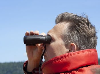 Side view of man looking through binoculars while standing against clear sky during sunny day