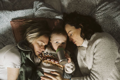 Girl drinking milk while lying amidst lesbian mothers on bed at home