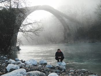 Full length of man crouching against arch bridge over river in foggy weather