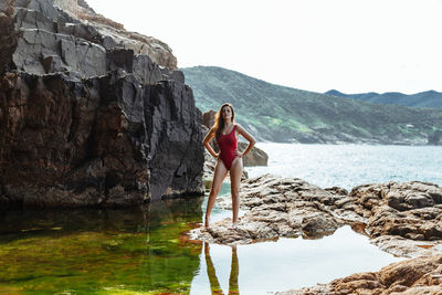 Portrait of woman standing on rock in sea against mountain and sky