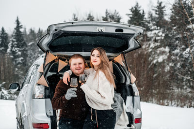 Couple holding wineglass while sitting by car trunk during winter