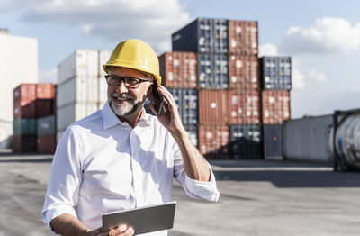 Businessman at cargo harbour, wearing safety helmet, using smartphone and digital tablet