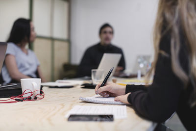 Midsection of young woman writing at desk while colleagues sitting in background