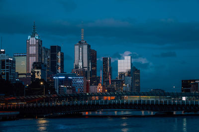 Illuminated buildings by river against sky in city