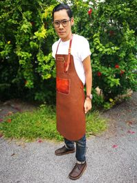 Full length portrait of man wearing apron while standing against plants on footpath