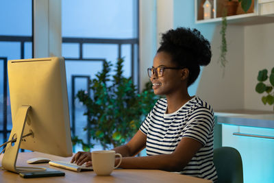 Young happy african woman remote worker wearing glasses typing on computer, enjoying freelance job