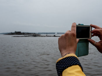 Cropped hands of woman photographing sea with smart phone against sky