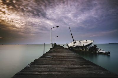 Pier by abandoned boat in sea against cloudy sky during sunset