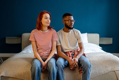 Pensive multiracial family couple sitting on comfortable bed daydreaming together looking to window.