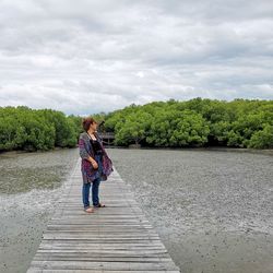 Full length of mid adult woman standing on boardwalk over lake against cloudy sky
