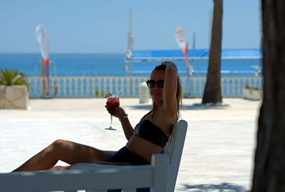 Young woman sitting on sunglasses at beach against sky