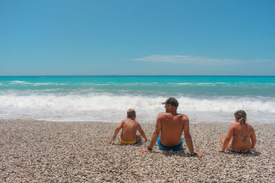 Rear view of shirtless man with children sitting at beach against blue sky