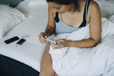 Woman looking at injection pen while sitting on bed in bedroom