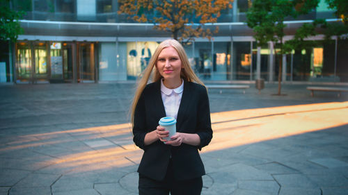 Portrait of young woman standing on footpath holding coffee cup