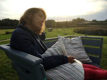 Woman sleeping on bench with cushions at park against sky during sunset