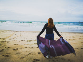 Rear view of woman with picnic blanket on beach against sky