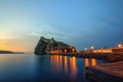 Illuminated buildings by sea against sky during sunset