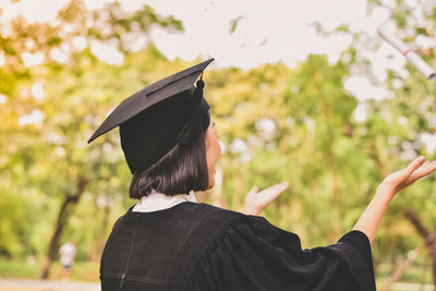 Happy young woman in graduation gown standing against trees