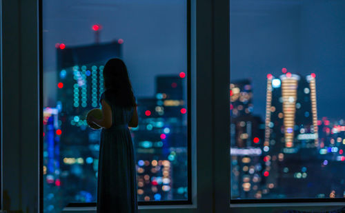 Rear view of woman standing by window at night