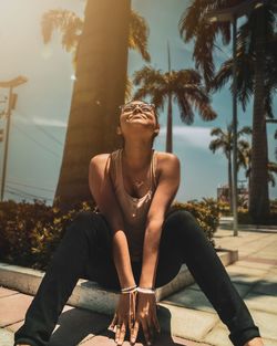 Woman looking up while sitting by palm tree on footpath