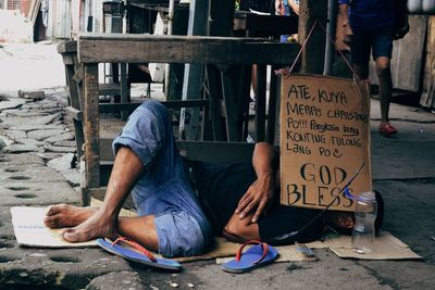 Homeless man lying on sidewalk by board with text