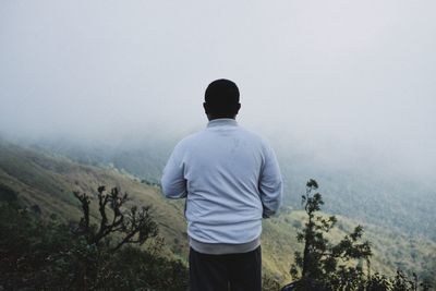 Rear view of man standing against mountain during foggy weather