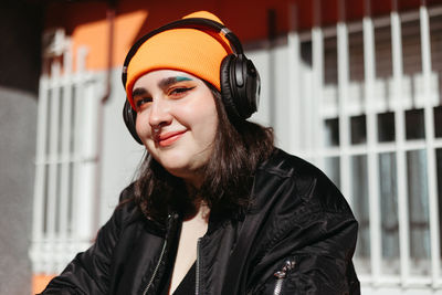 Female listening to music in wireless headphones while standing on street near shabby wall looking at camera