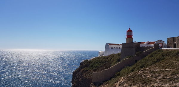 Lighthouse by sea against buildings against clear blue sky