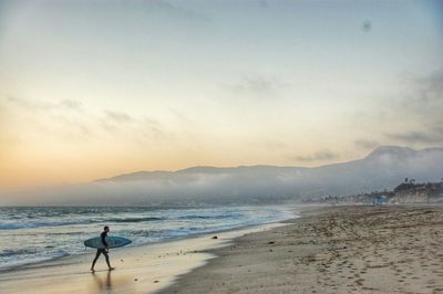 Man carrying surfboard at beach against sky